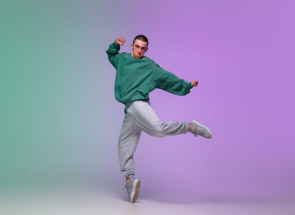 Boy dancing hip-hop in stylish clothes on gradient background at dance hall in neon light.